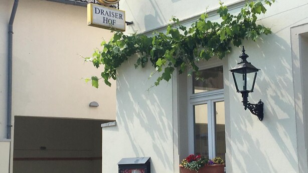 A building with a sign reading "DRAISER HOF" and a clock above it. There is a window with a flower box and a wall-mounted light fixture.