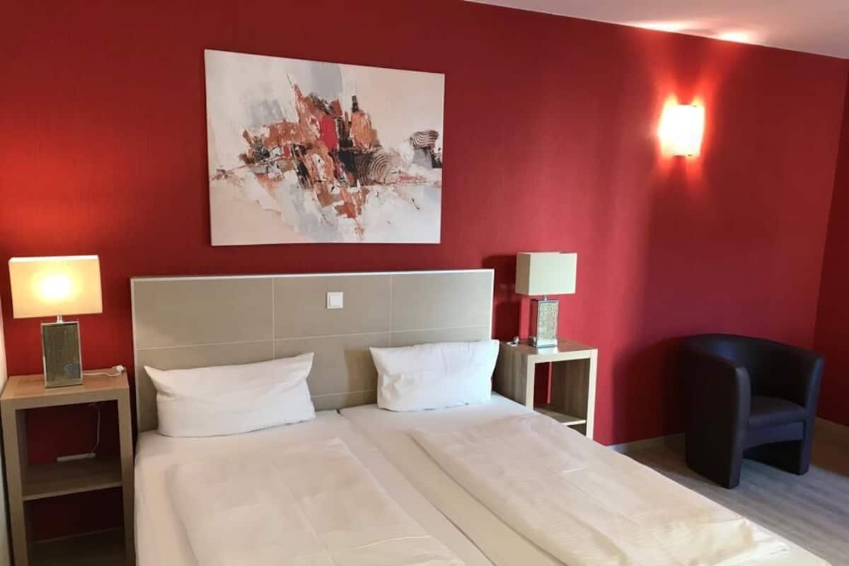 Habitación de hotel con una cama grande, dos mesitas de noche con lámparas, una silla y una pared roja decorada con un cuadro.