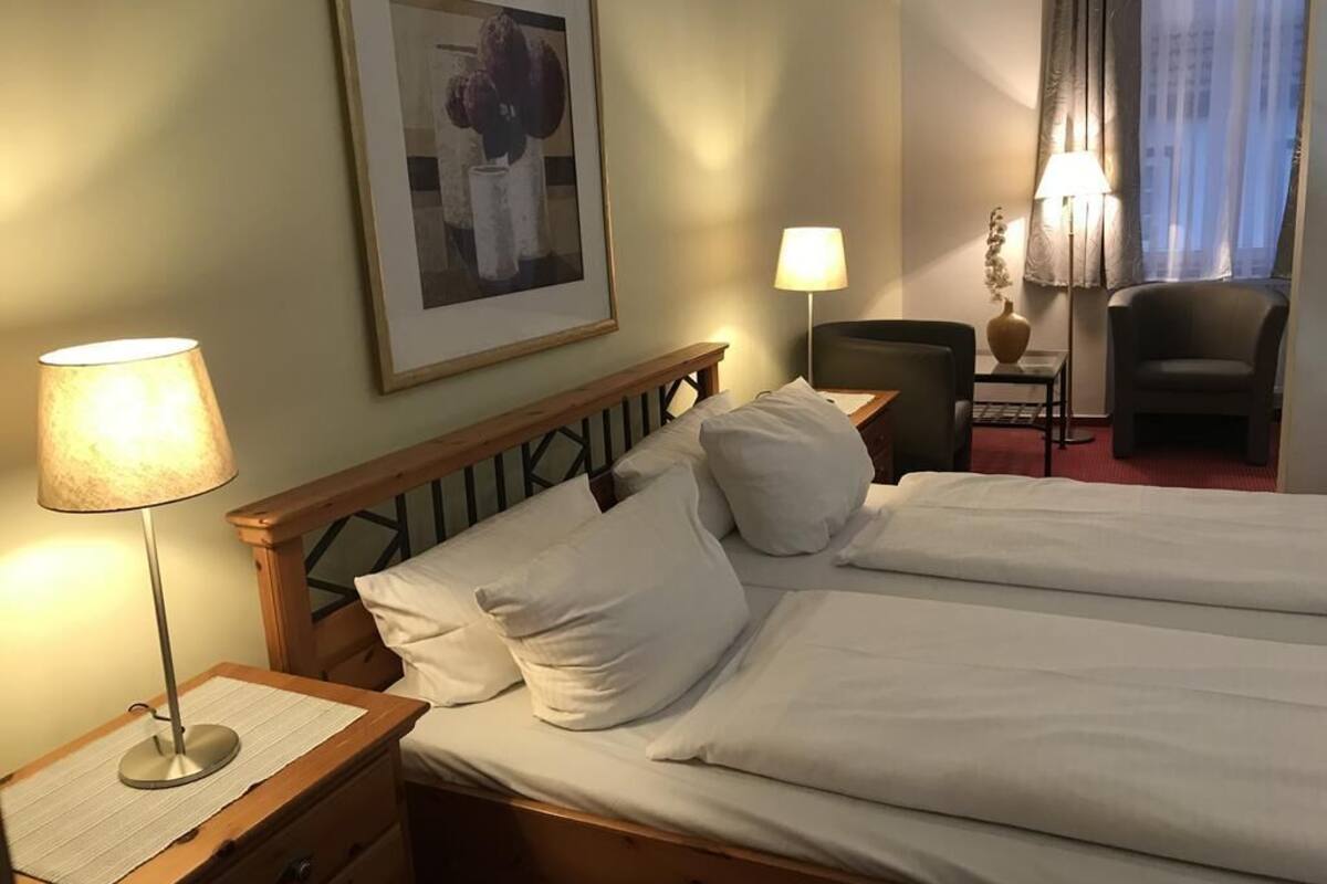 Habitación de hotel con dos camas, una mesita de noche con lámpara, una silla, una pequeña mesa con lámpara y un cuadro enmarcado en la pared.