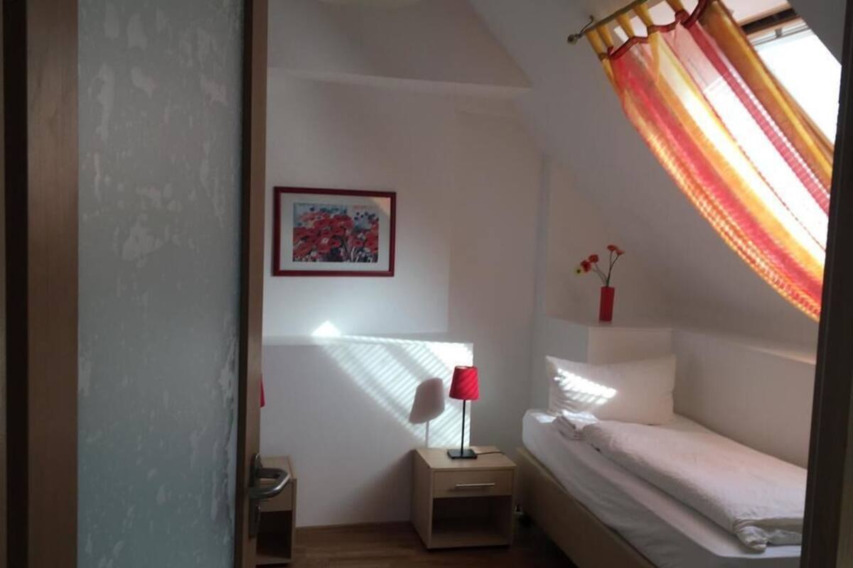 Una habitación pequeña y luminosa con una cama individual, una mesita de noche con lámpara, un cuadro enmarcado en la pared y una ventana con cortinas.