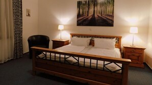 Ein Hotelzimmer mit einem hölzernen Bett, zwei Nachttischen mit Lampen, einem Sessel und einem Waldgemälde an der Wand.