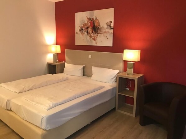 A double bed with white linens, two bedside tables with lamps, a red accent wall, and a painting above the bed.