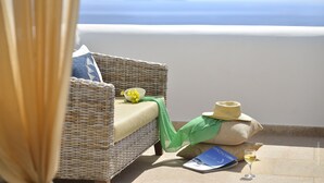 Une chaise en osier avec un coussin, un bol de raisins, un chapeau de paille, un livre et un verre de vin sur un balcon donnant sur la mer.