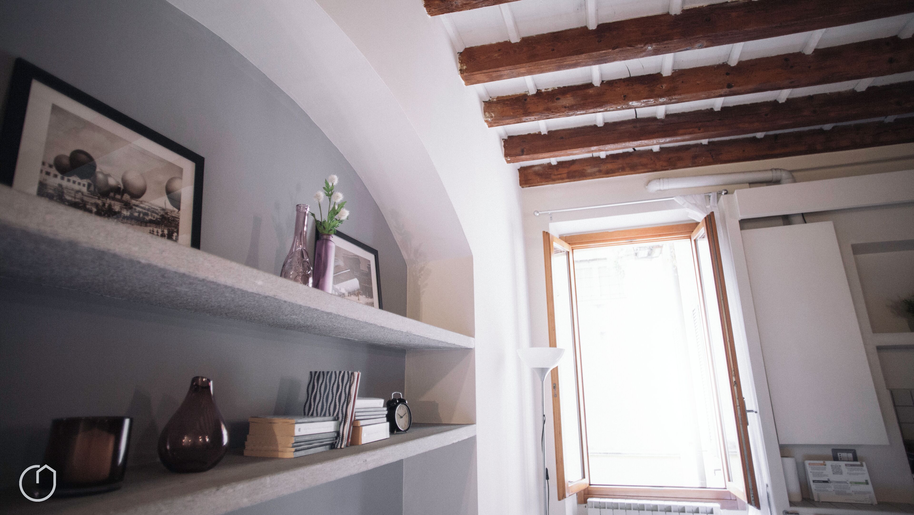 Una stanza con soffitto spiovente, travi in legno, una finestra con tende e scaffali contenenti diversi oggetti, tra cui libri, un vaso e un quadro incorniciato.