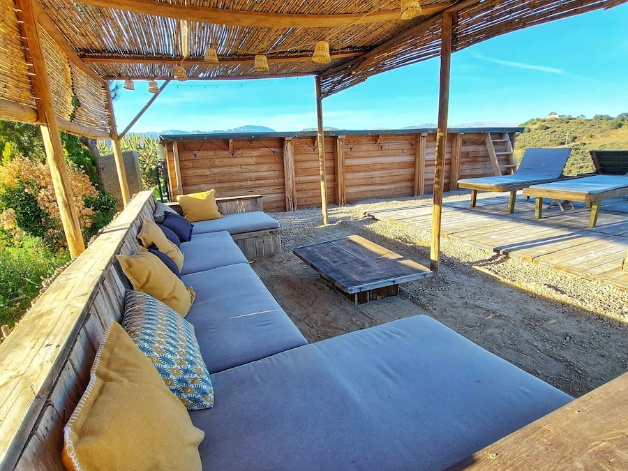 An outdoor seating area with a wooden bench and cushions, a wooden table, and a bamboo ceiling.