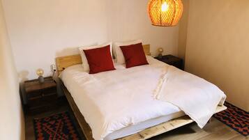 A bed with white bedding and two red pillows, a wooden headboard, a bedside table with a lamp, and a hanging light fixture.