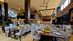 A spacious dining area with multiple tables set for a meal, a person serving food, and a view of the upper floor. - HOTEL GRAND BARON TAIPNG (TAIPING)