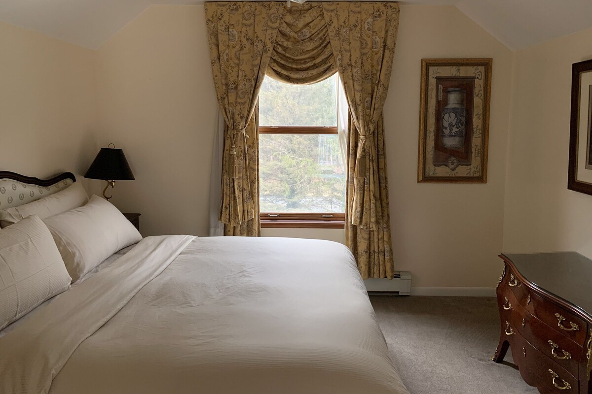 A bedroom with a bed, a ceiling fan, a window with curtains, a bedside lamp, a dresser, and a framed picture on the wall.