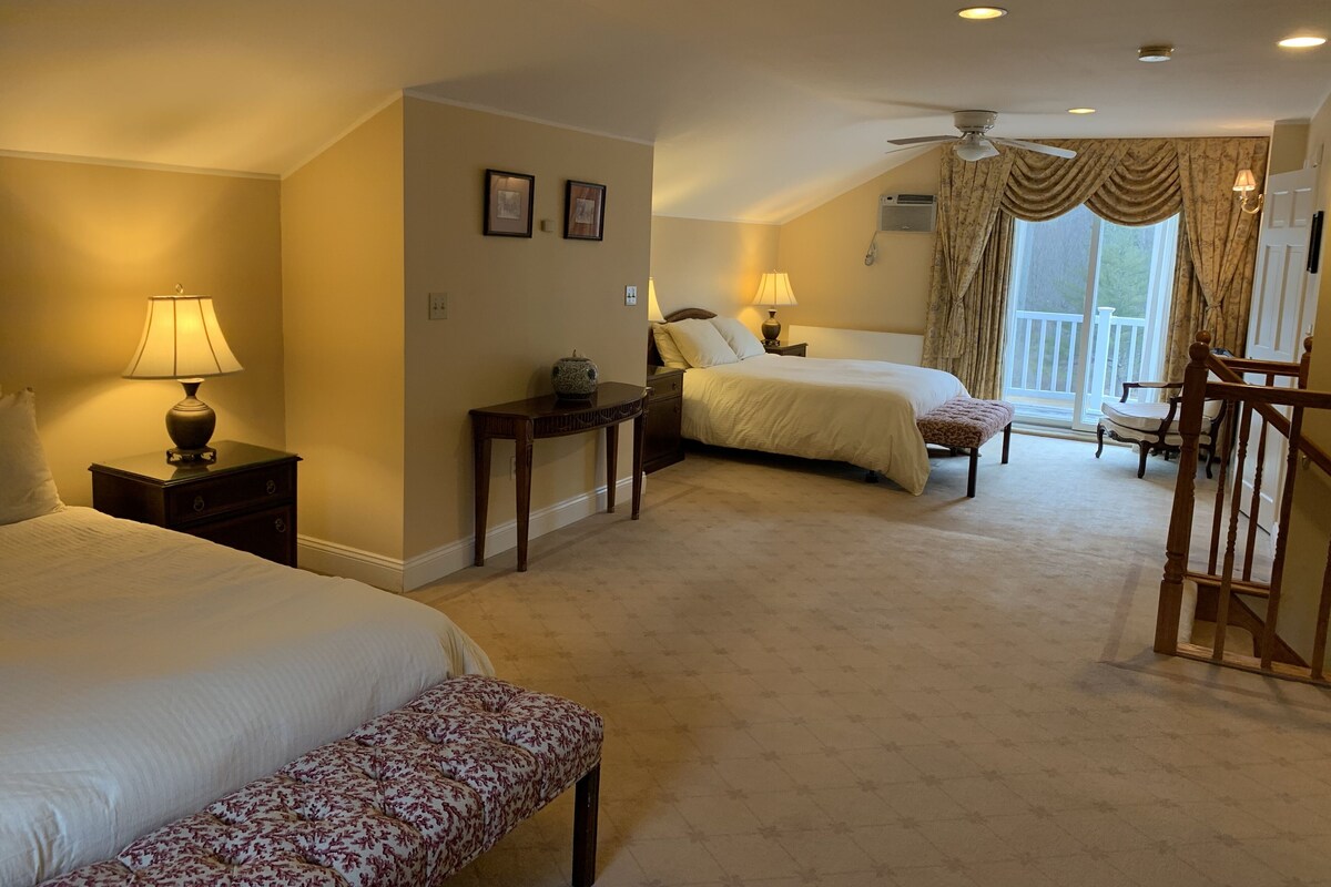 A hotel room with two beds, a desk, a chair, a bench, and a balcony with a view.