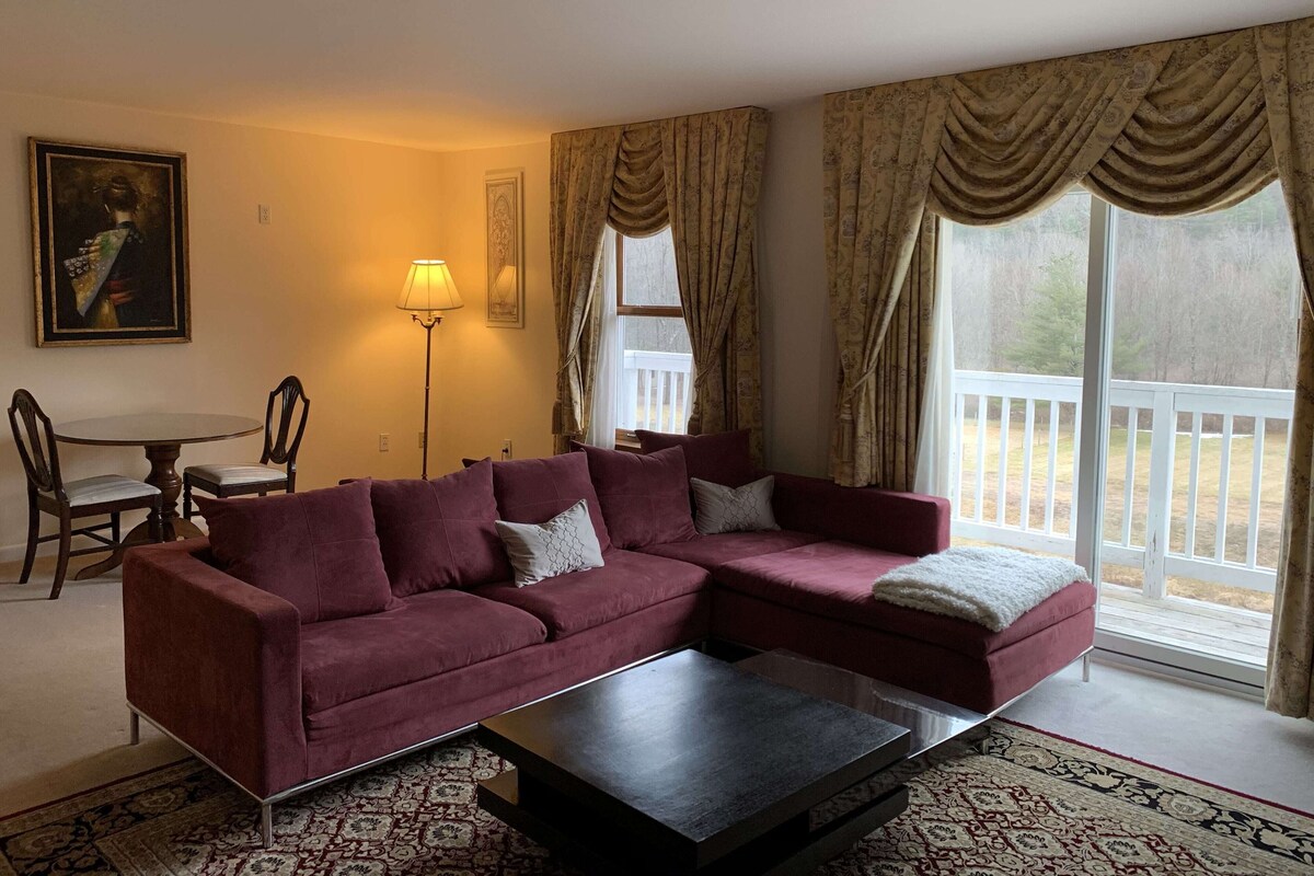 A living room with a sectional sofa, a coffee table, a lamp, a dining table with chairs, and a view of a deck through large windows.