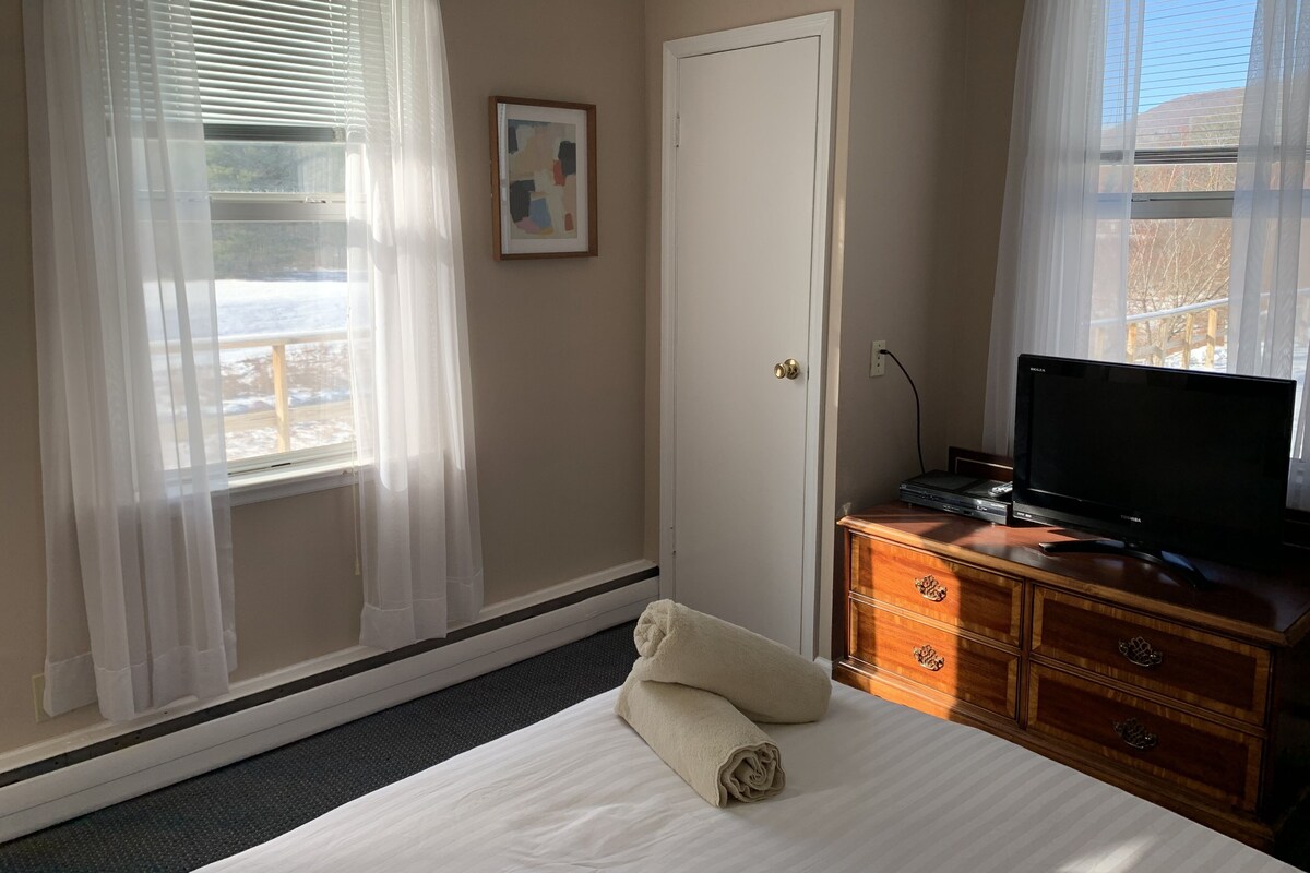 A hotel room with a bed, a television on a wooden dresser, a door, and a window with sheer curtains.