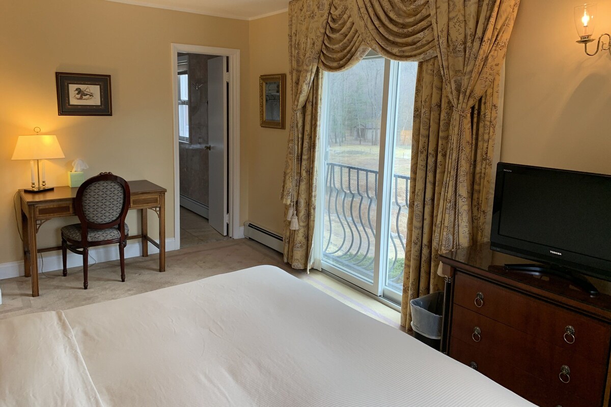 A hotel room with a bed, a desk with a chair, a television, and a balcony with curtains.