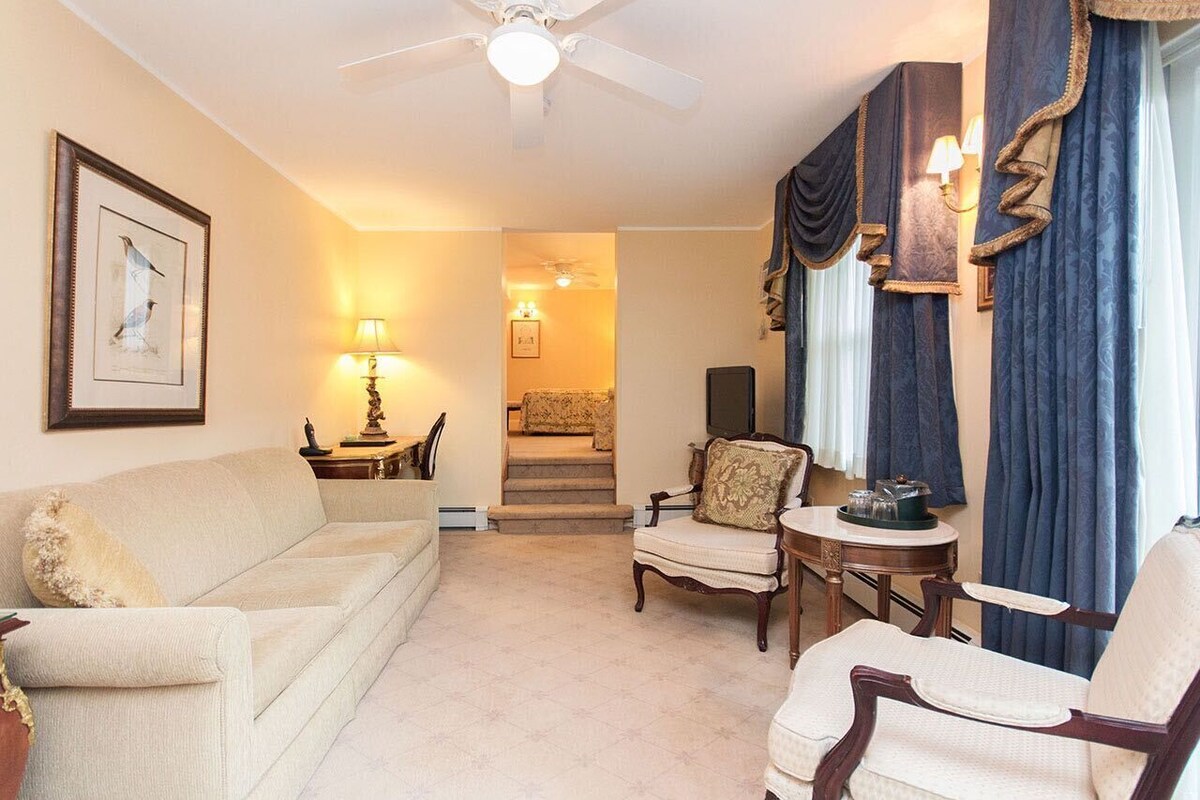 A cozy living room with a sofa, armchairs, a small table, and a ceiling fan. A staircase leads to another room.