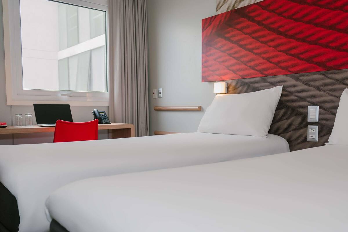 A hotel room with two beds, a desk with a laptop and phone, and a window with curtains.