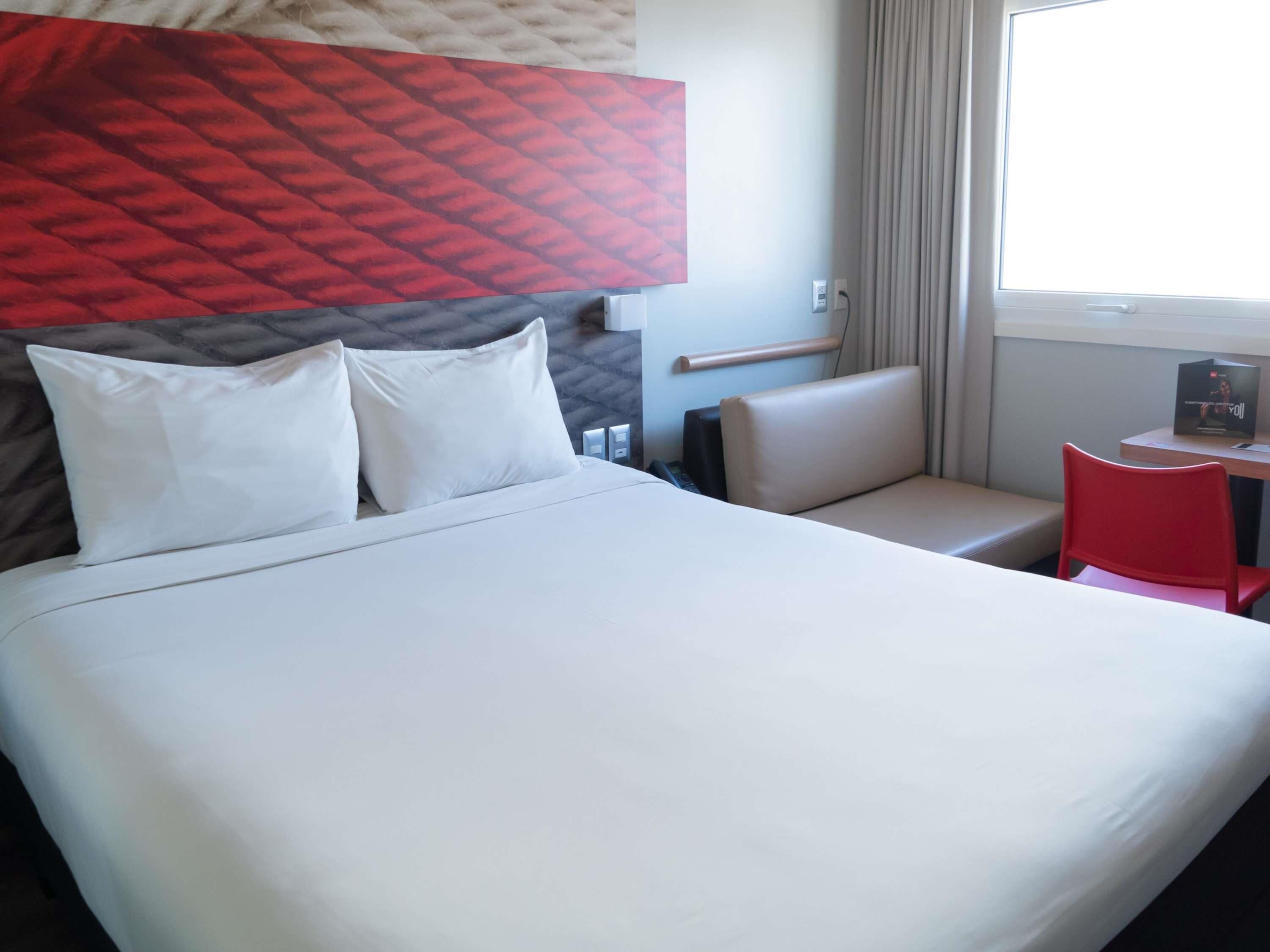 A hotel room with a large bed, a red chair, a small table, and a window with curtains.