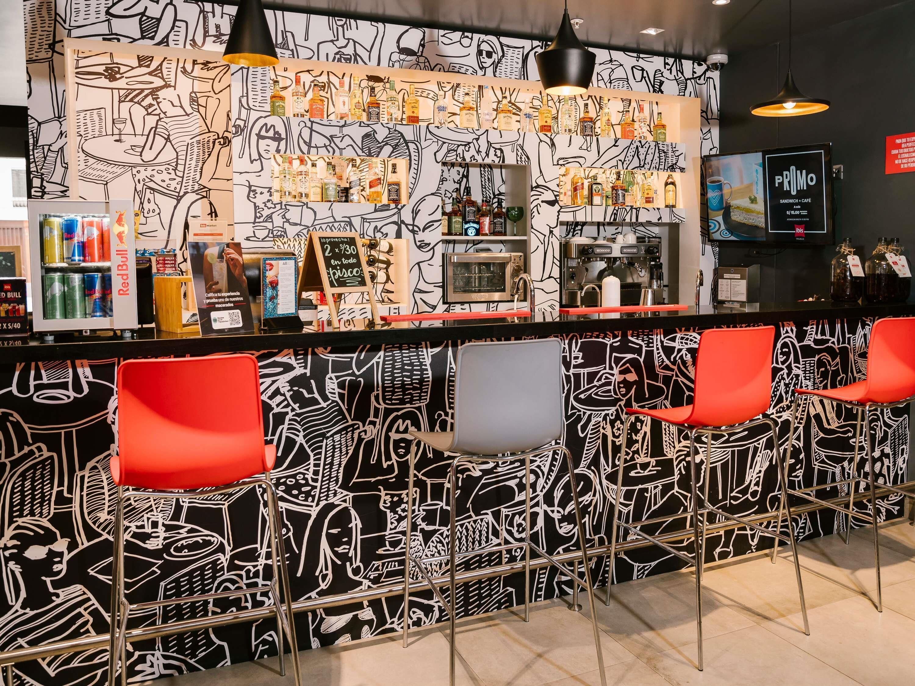 Zona de bar con un mural en blanco y negro, sillas rojas, una barra con bebidas y una máquina de café.