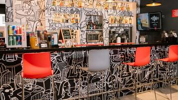 Zona de bar con un mural en blanco y negro, sillas rojas, una barra con bebidas y una máquina de café.