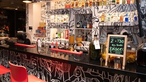 A bar area with a black countertop, red bar stools, and a wall decorated with black and white drawings. There are shelves with bottles and a chalkboard sign advertising a promotion. - ibis Trujillo (Trujillo)