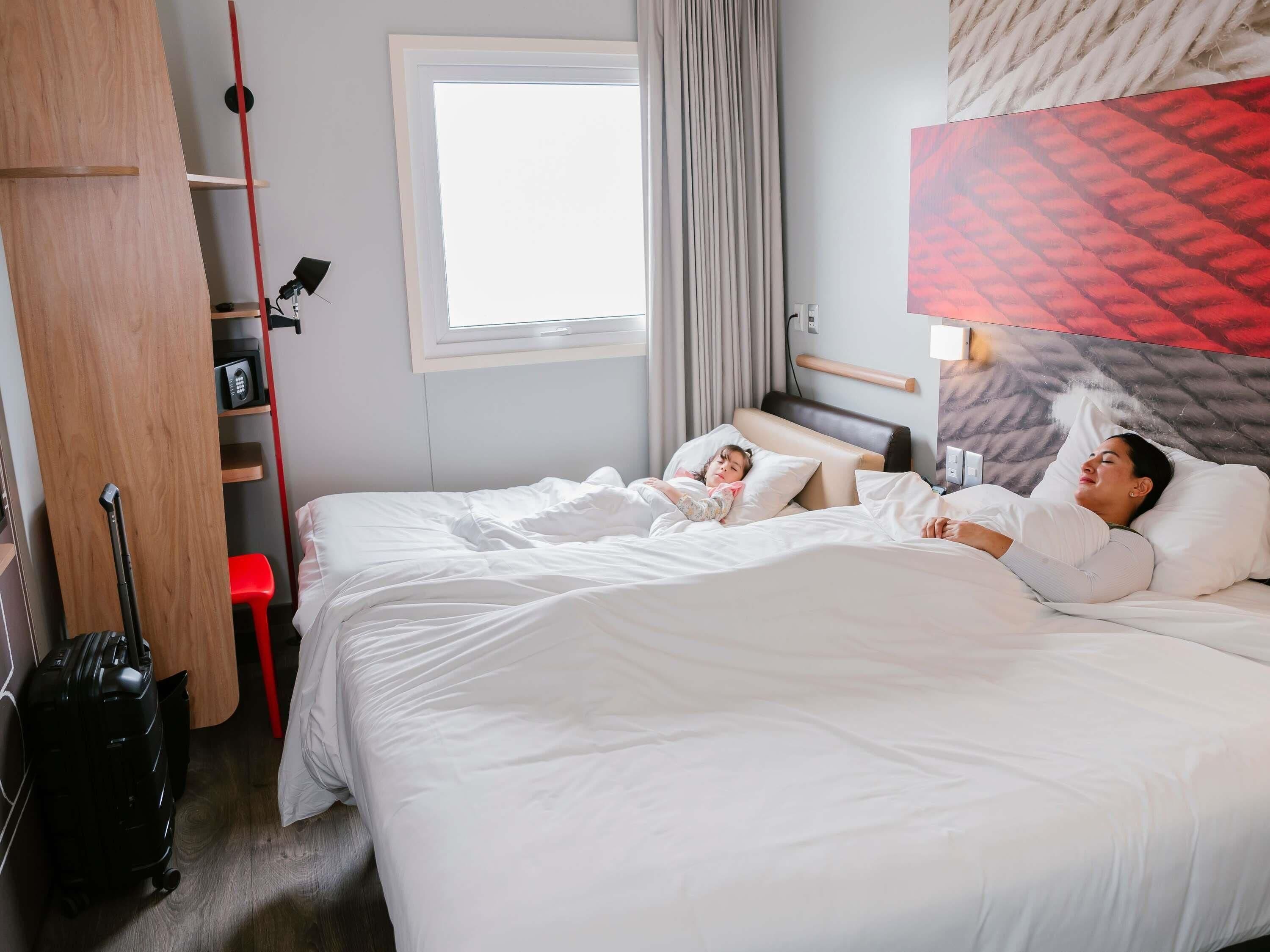 Et hotelværelse med en seng, en kuffert, en rød skammel, et vindue og en vægmonteret lampe.