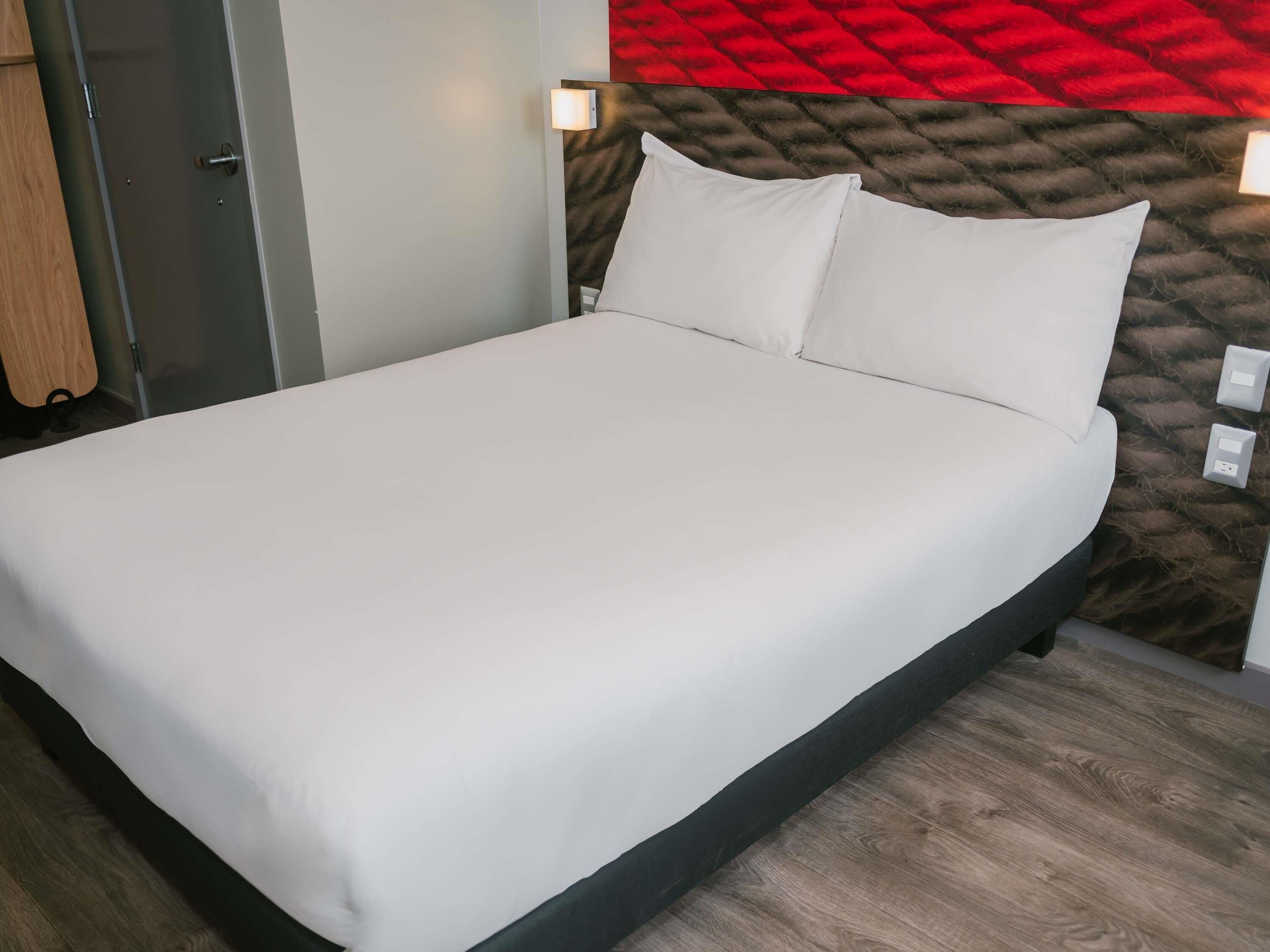 Een bed met witte lakens, een hoofdbord met een rood en bruin patroon, en een houten vloer.