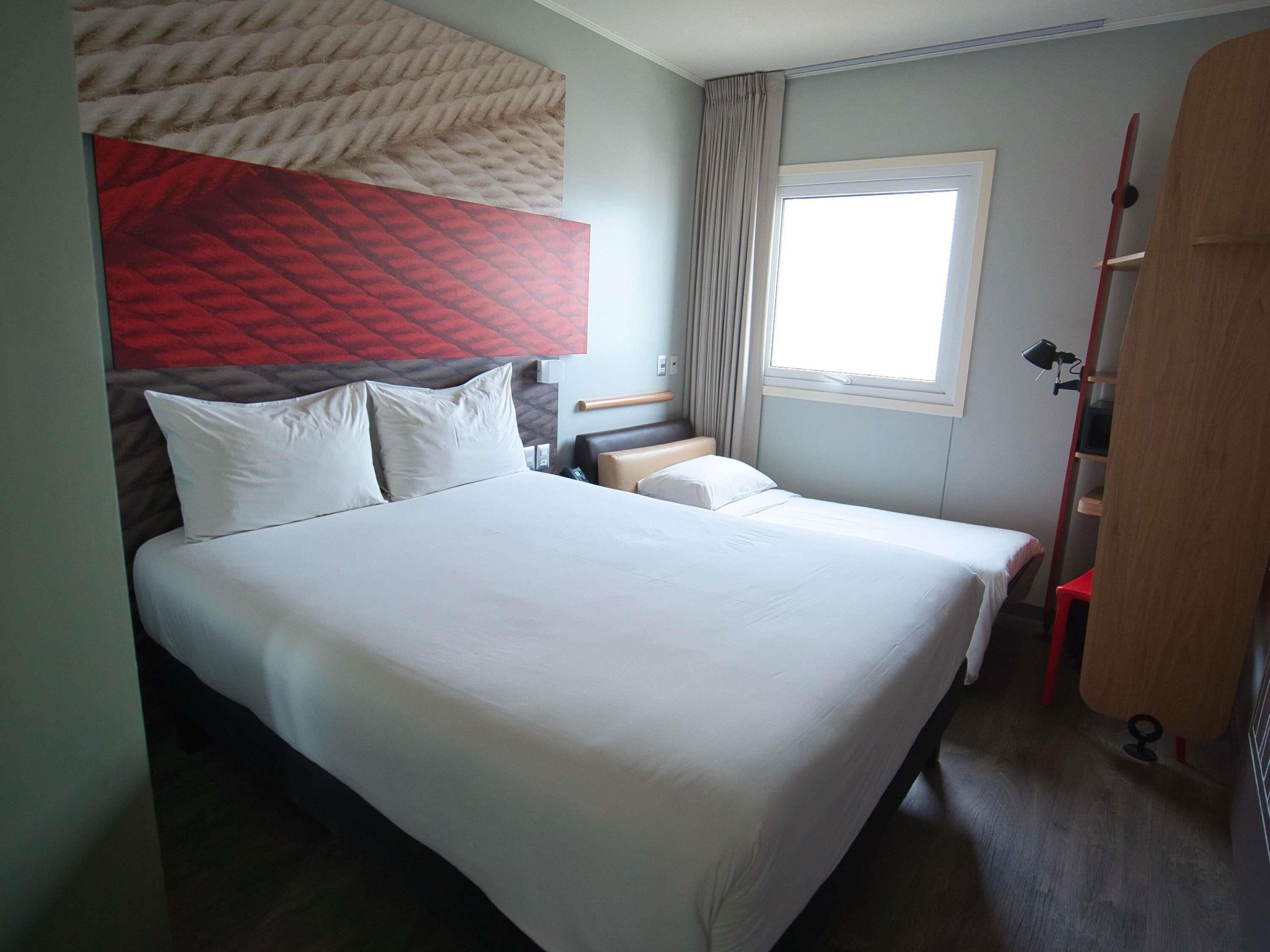 Habitación de hotel con una cama grande, otra más pequeña, una ventana con cortinas, una puerta de madera y un cabecero con estampado rojo y beige.