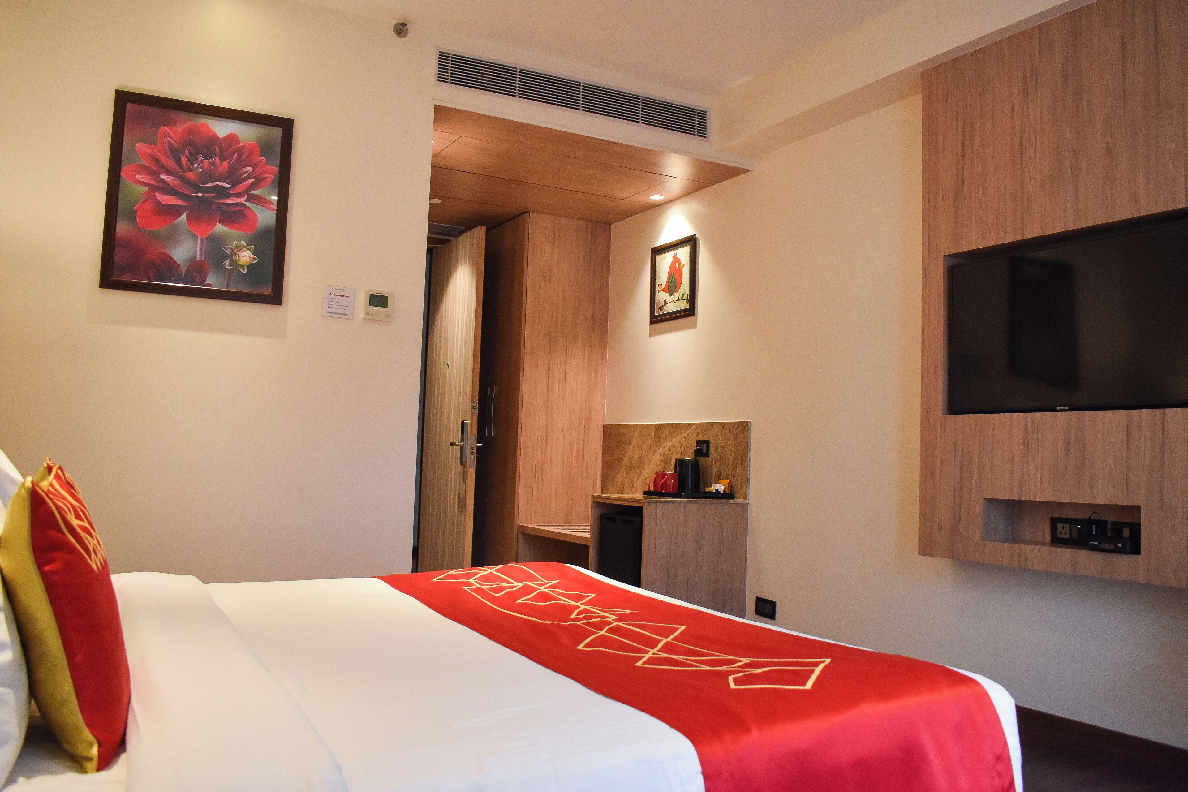 A hotel room with a bed, a TV mounted on the wall, a wooden wardrobe, and a framed picture on the wall.