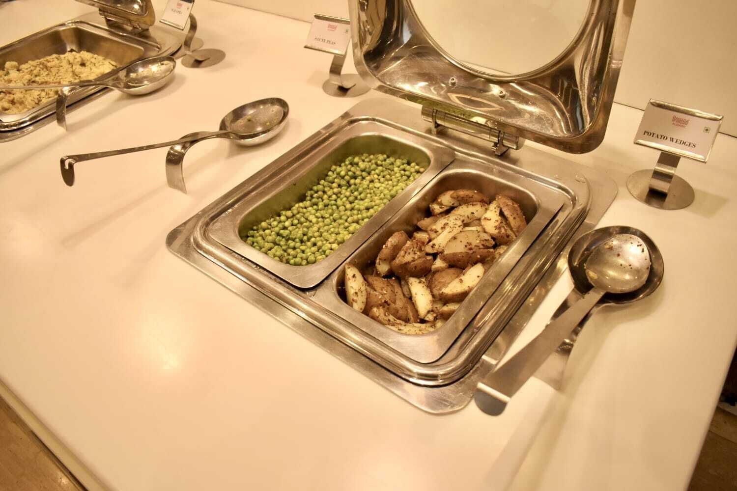 A buffet with labeled containers of potato wedges and green peas.
