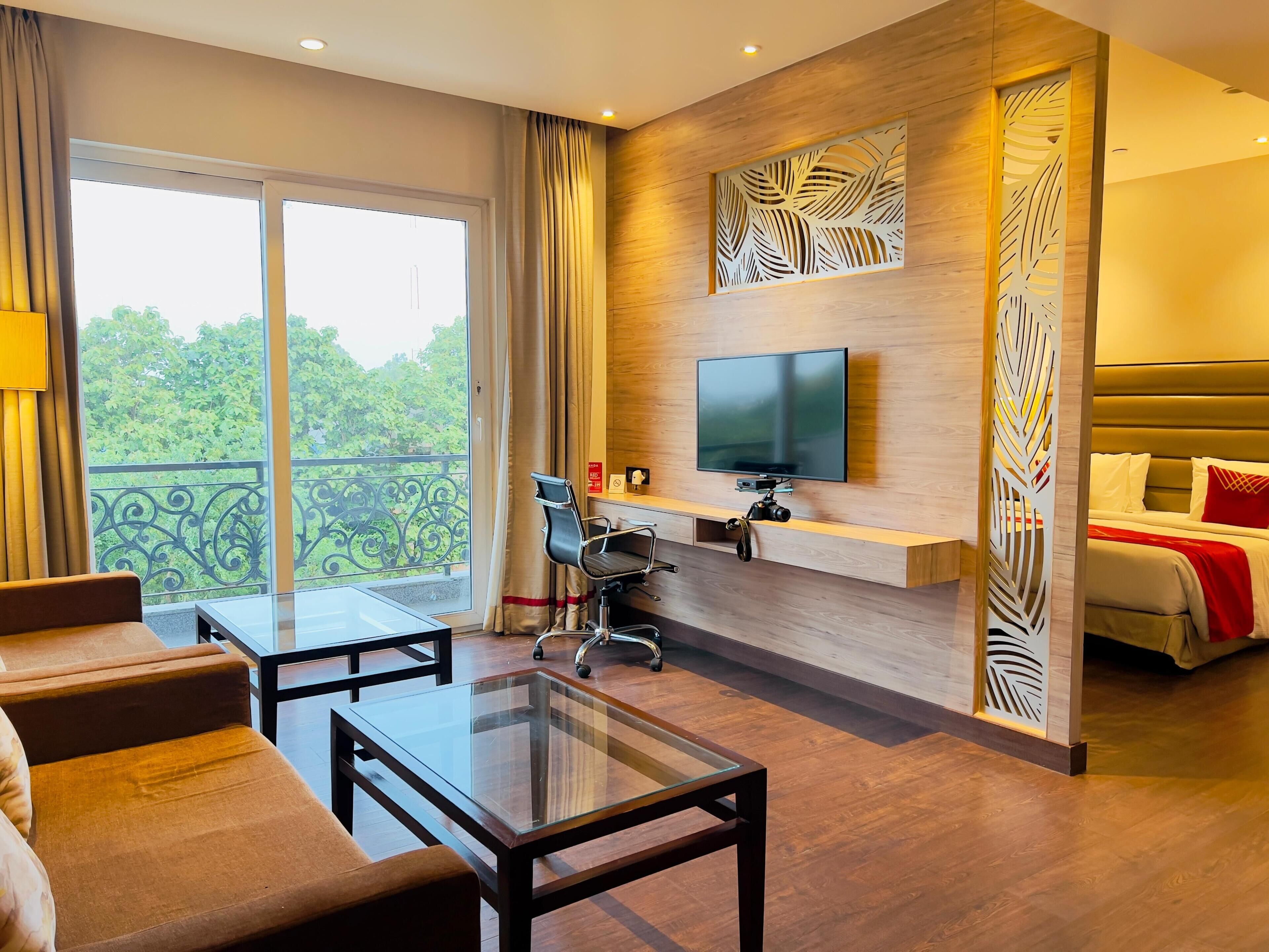 A modern hotel room with a large balcony, a flat-screen TV mounted on a wooden panel, a sofa, a glass-top coffee table, and a bed with red pillows.