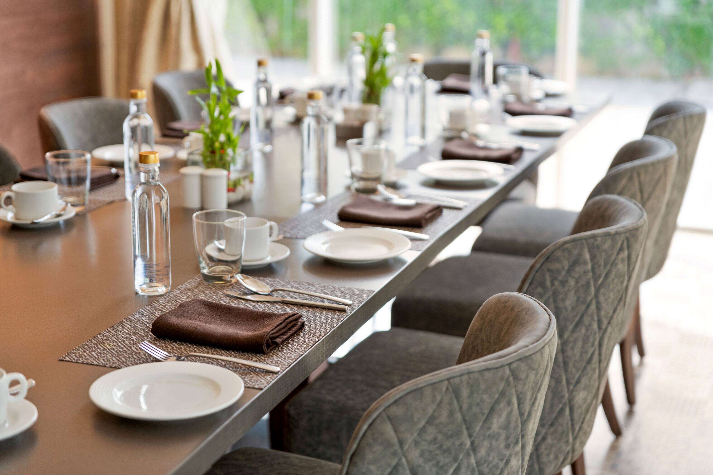 A long dining table set with plates, glasses, cutlery, and napkins, surrounded by upholstered chairs.