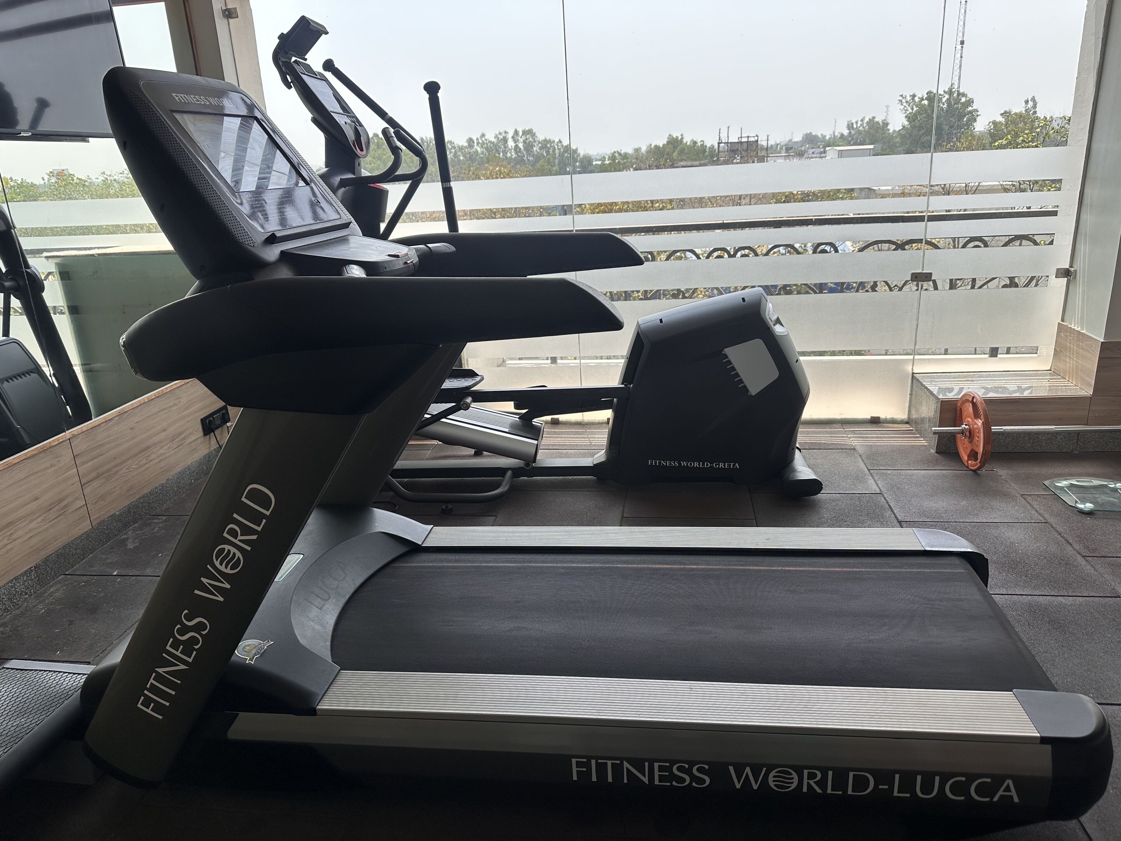 A treadmill labeled 'FITNESS WORLD' and 'FITNESS WORLD-LUCCA' in a gym with a view of a body of water through large windows.