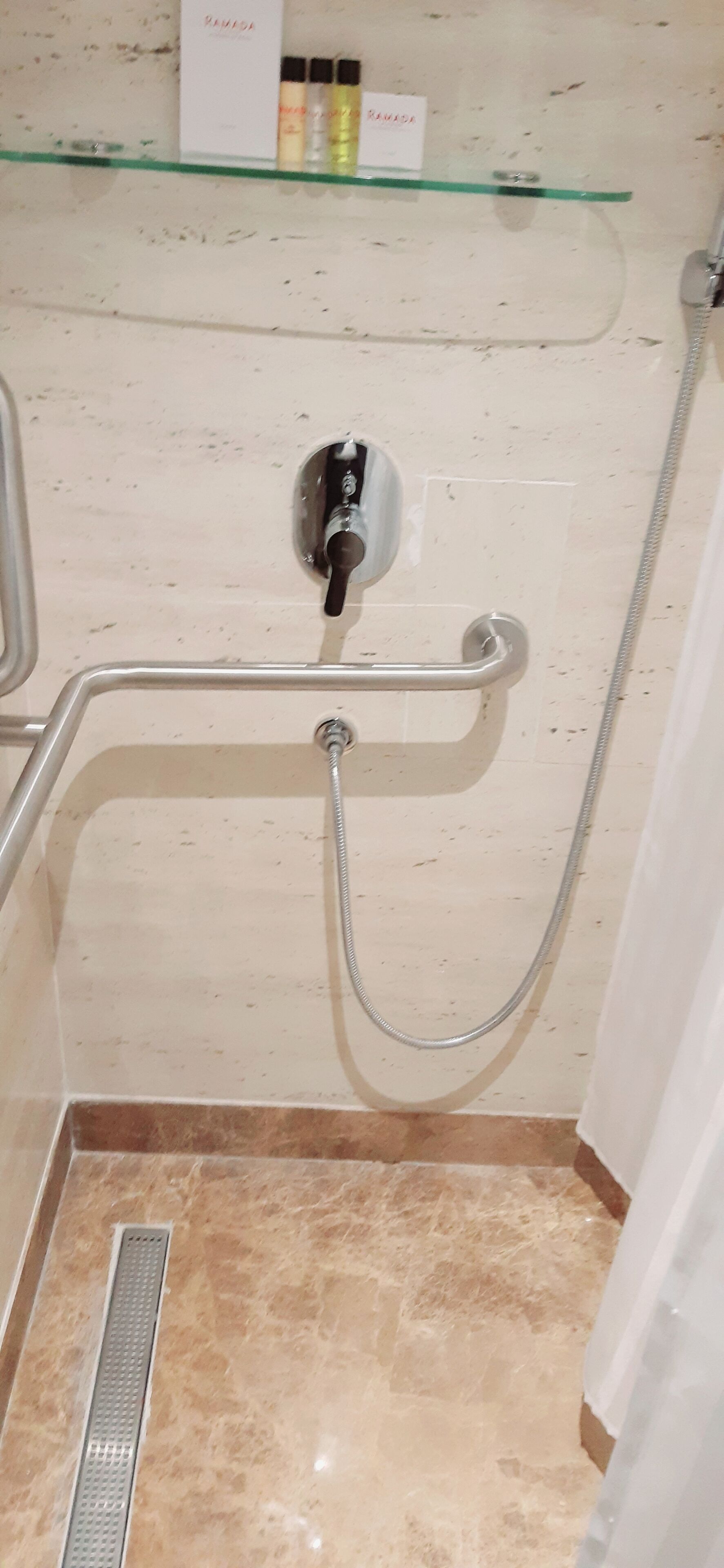 A bathroom with a glass shelf holding toiletries, a handheld showerhead, and a tiled floor.