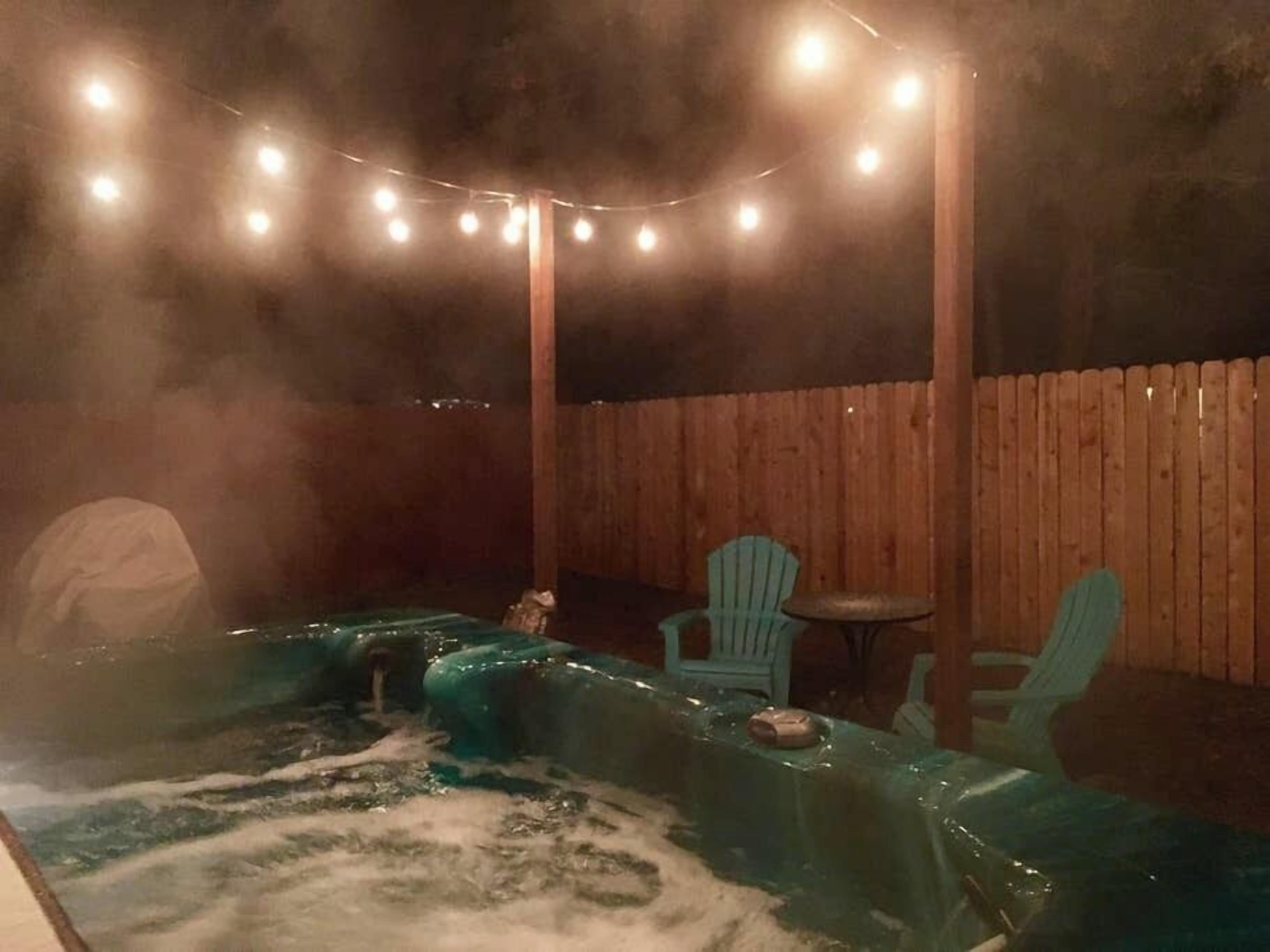 Area con idromassaggio, recinzione in legno, luci a stringa e due sedie verdi.