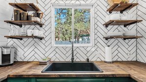 A kitchen with a herringbone tile backsplash, a window with a view of trees, and wooden shelves with kitchen items.
