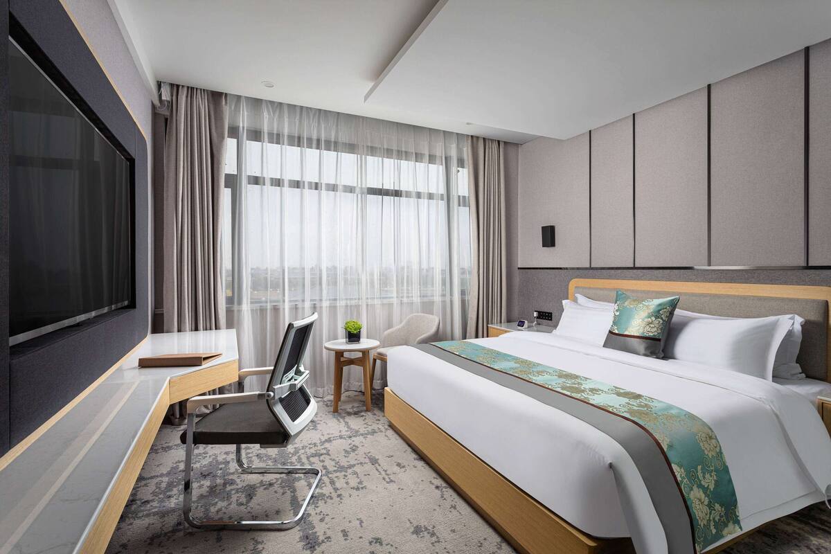 A modern hotel room with a large bed, a desk, a TV, and a view of the city through large windows.