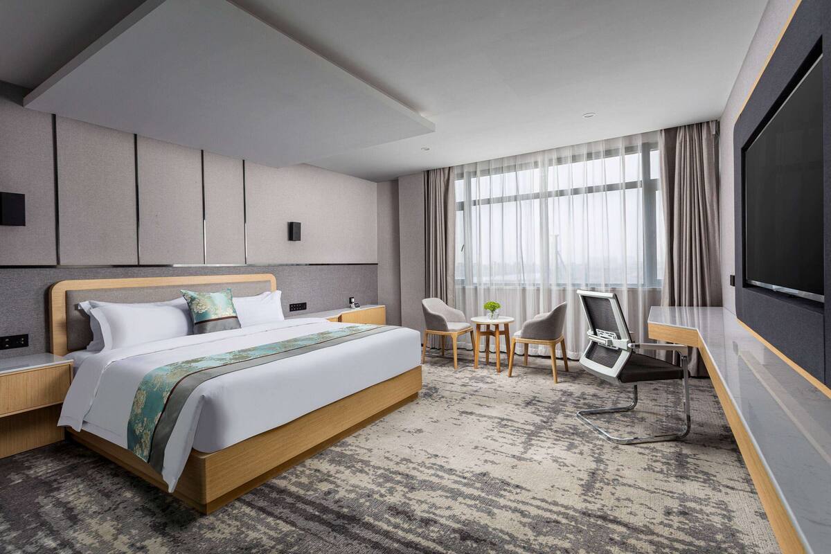 A modern hotel room with a large bed, a desk, two chairs, a TV, and a large window with curtains.