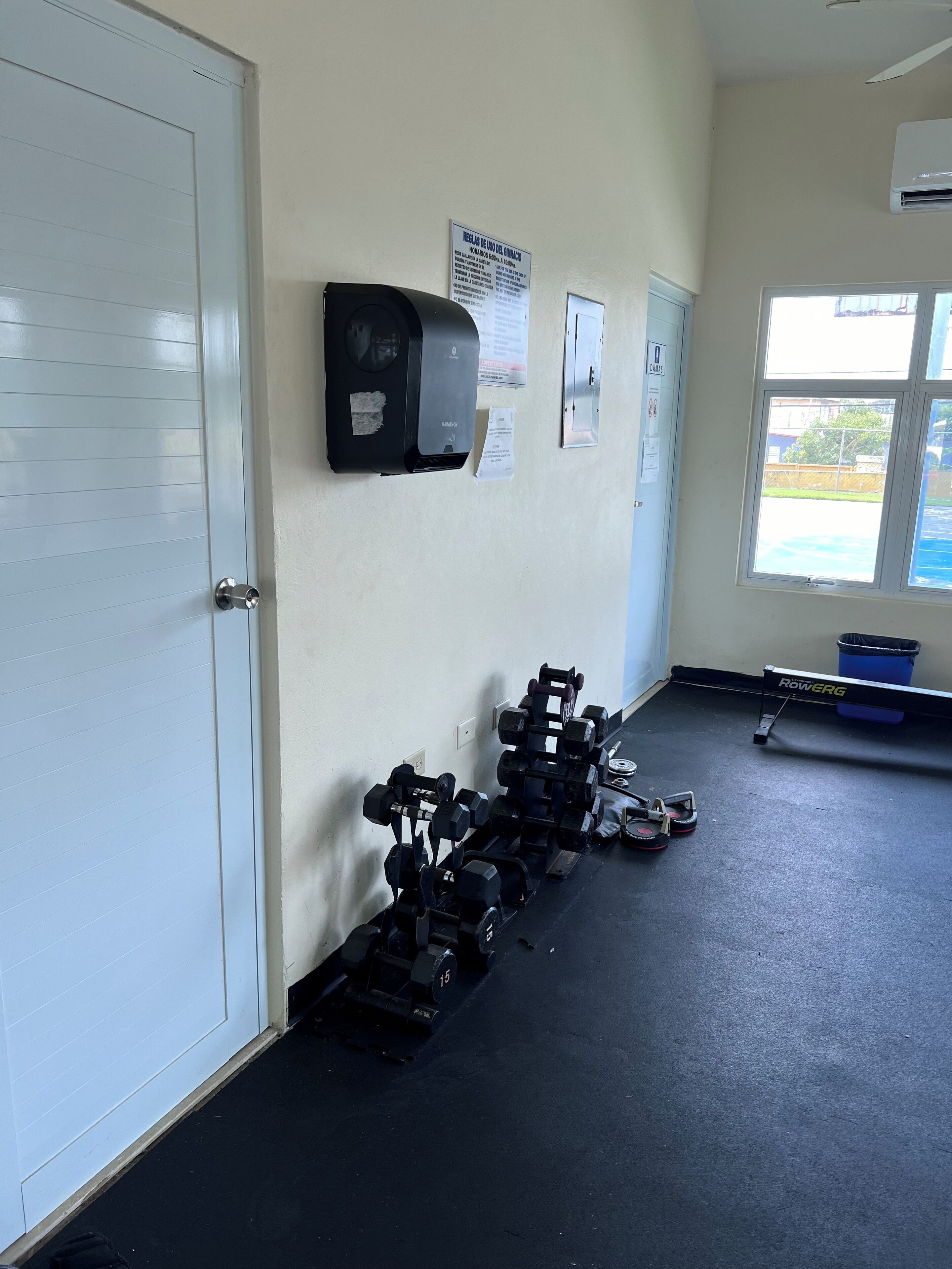 A gym with weightlifting equipment, a wall-mounted air conditioner, and a window with a view of the outside.