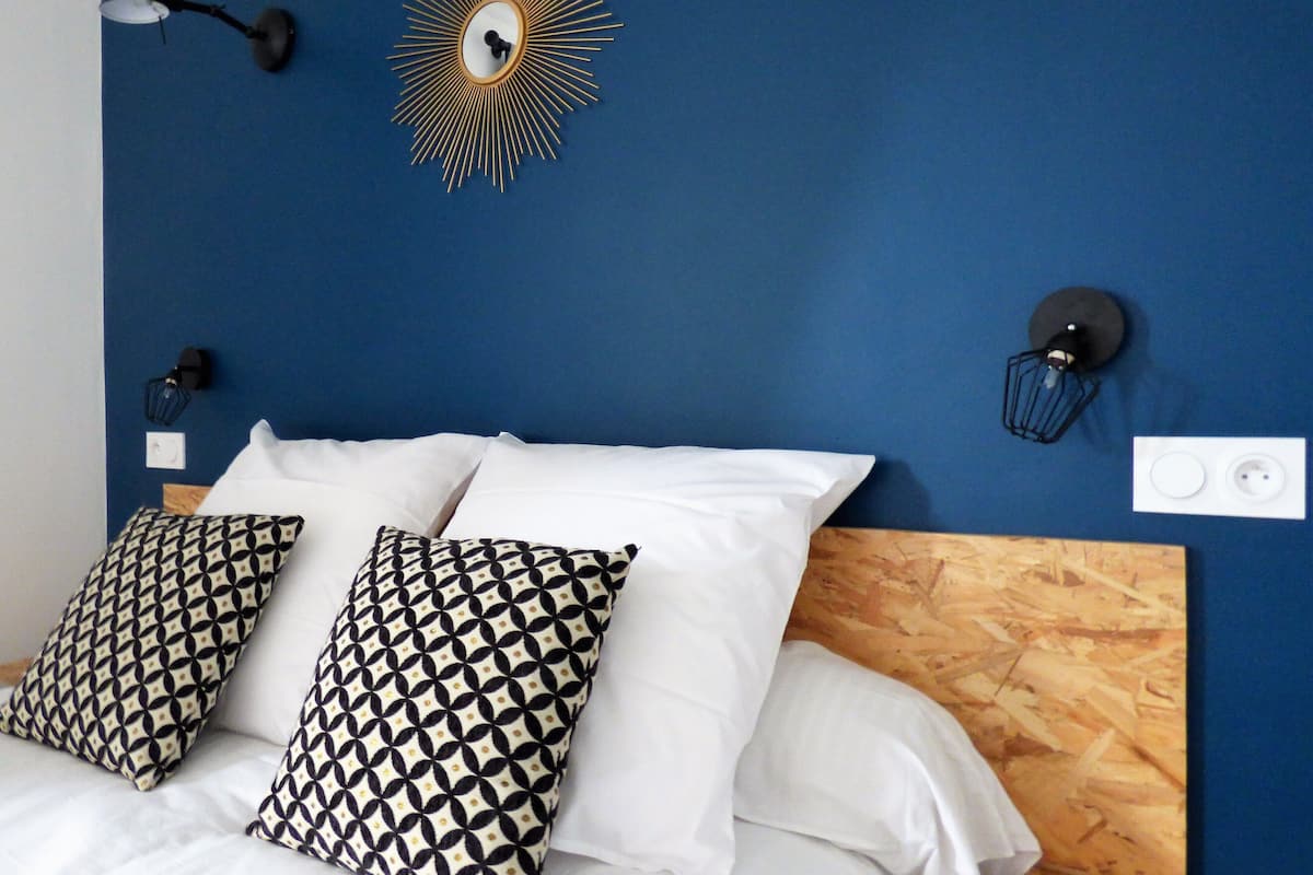 Un lit bien fait, avec du linge de lit blanc et des coussins à motifs, une tête de lit en bois et un mur bleu orné d’un miroir en forme de soleil et d’appliques murales.