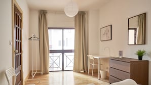 A hotel room with a wooden desk, a white chair, a bed with white linens, a hanging light fixture, and a view through a large window with curtains. - Campo Pequeno Avenida (Lisbon)