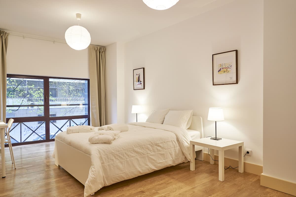Een slaapkamer met een groot bed, twee bedlampen, een raam met gordijnen en twee ingelijste foto’s aan de muur.