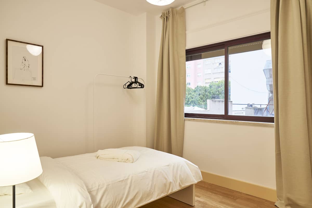 Een klein, netjes opgemaakt bed in een kamer met een raam, een wandlamp en een lijst met een afbeelding.