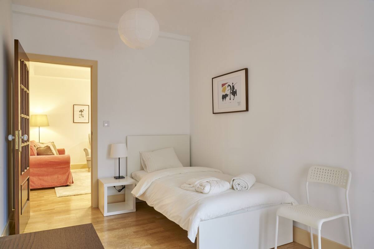 Een slaapkamer met een bed, nachtkastje, stoel, lamp en een deur naar een andere kamer.