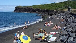 A beach with people sunbathing and swimming, a rocky shoreline, and a small structure in the background. - Great Tranquility Holiday Home (S.Roque do Pico)