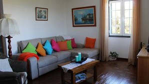 A living room with a grey sofa, colorful cushions, a wooden coffee table with magazines, a lamp, and two paintings on the wall. - Great Tranquility Holiday Home (S.Roque do Pico)