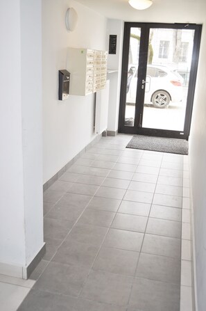 A hallway with a tiled floor, a door with a mat, and a wall-mounted light fixture.