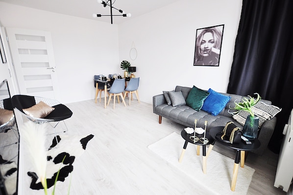 A modern living room with a grey sofa, black and white patterned pillows, a black coffee table, and a dining area with a black table and blue chairs.