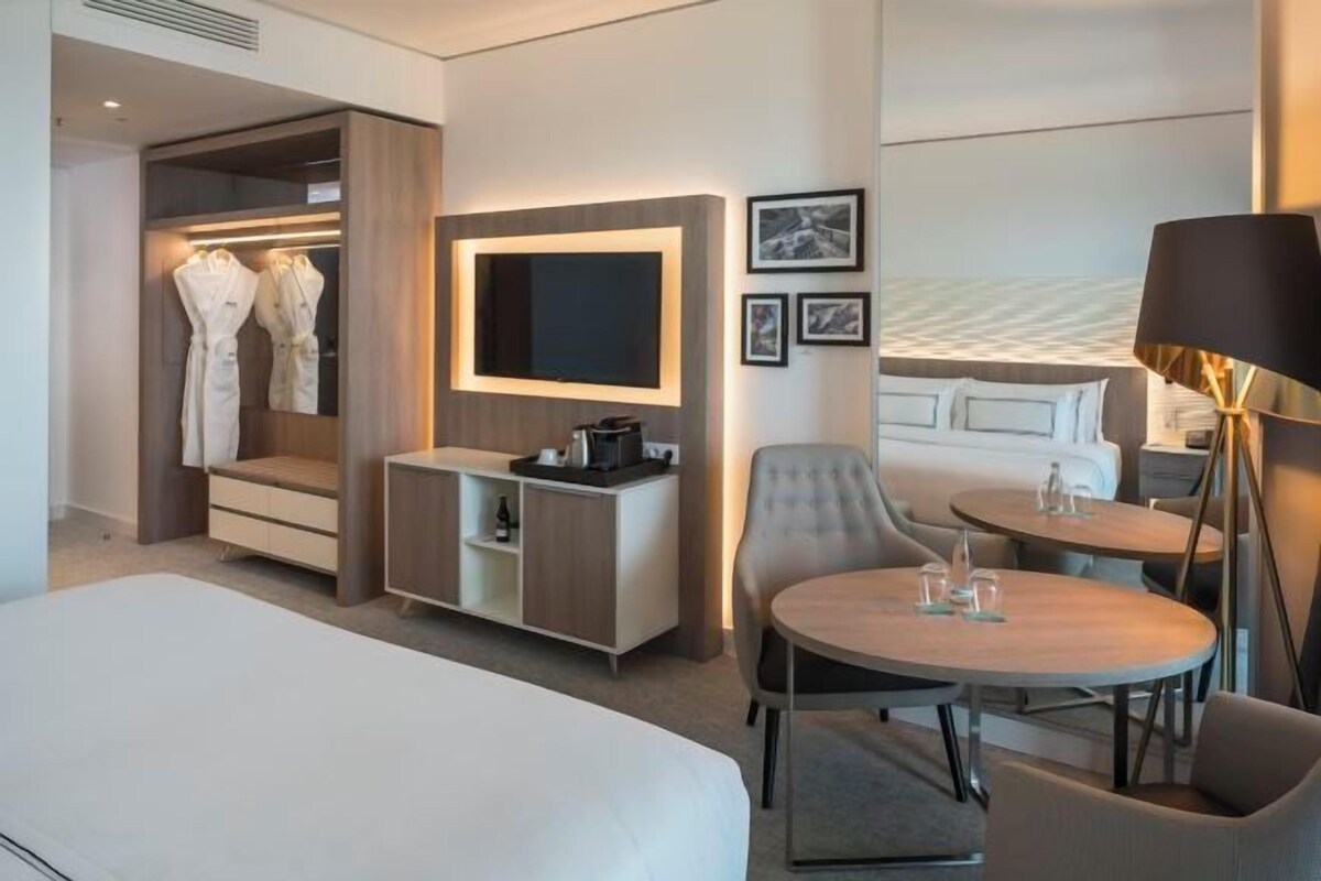 A modern hotel room with a bed, a seating area with a table and chairs, a TV mounted on the wall, a wardrobe with robes, and a bedside table with a lamp.