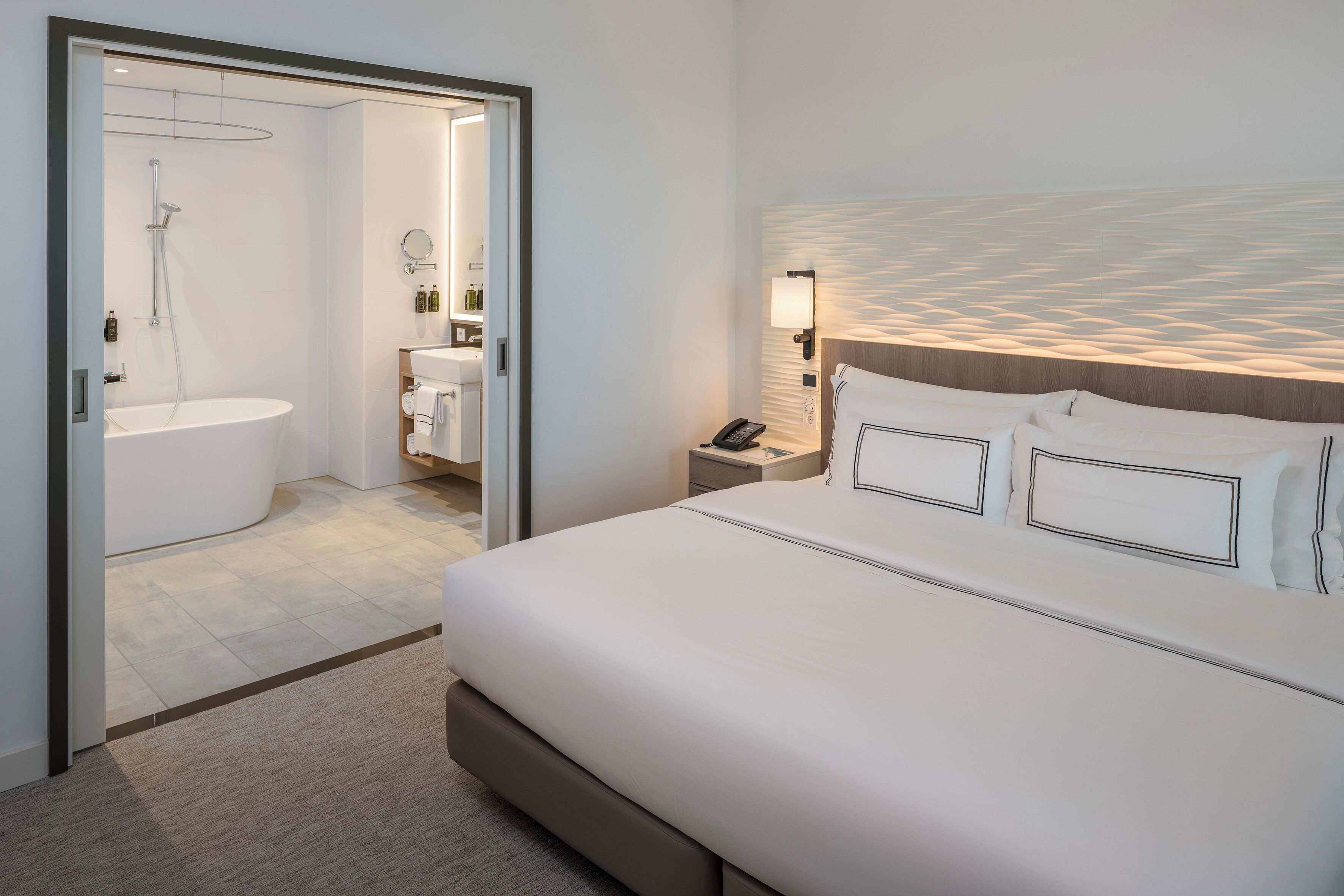 Ein modernes Hotelzimmer mit einem großen Bett, einem Nachttisch mit Lampe, einem Badezimmer mit Dusche und Waschbecken sowie Blick auf den Strand.