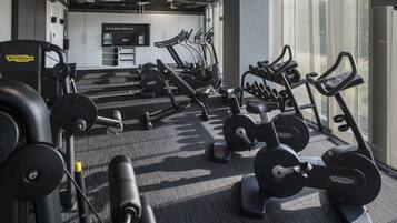 A gym with various exercise machines, a wall-mounted TV, and a window providing natural light.