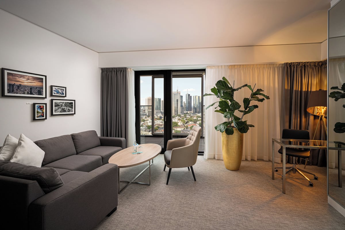Ein modernes Hotelzimmer mit einem grauen Sofa, einem kleinen runden Tisch, einem Sessel, einer großen Topfpflanze und Blick auf die Stadt durch das Fenster.