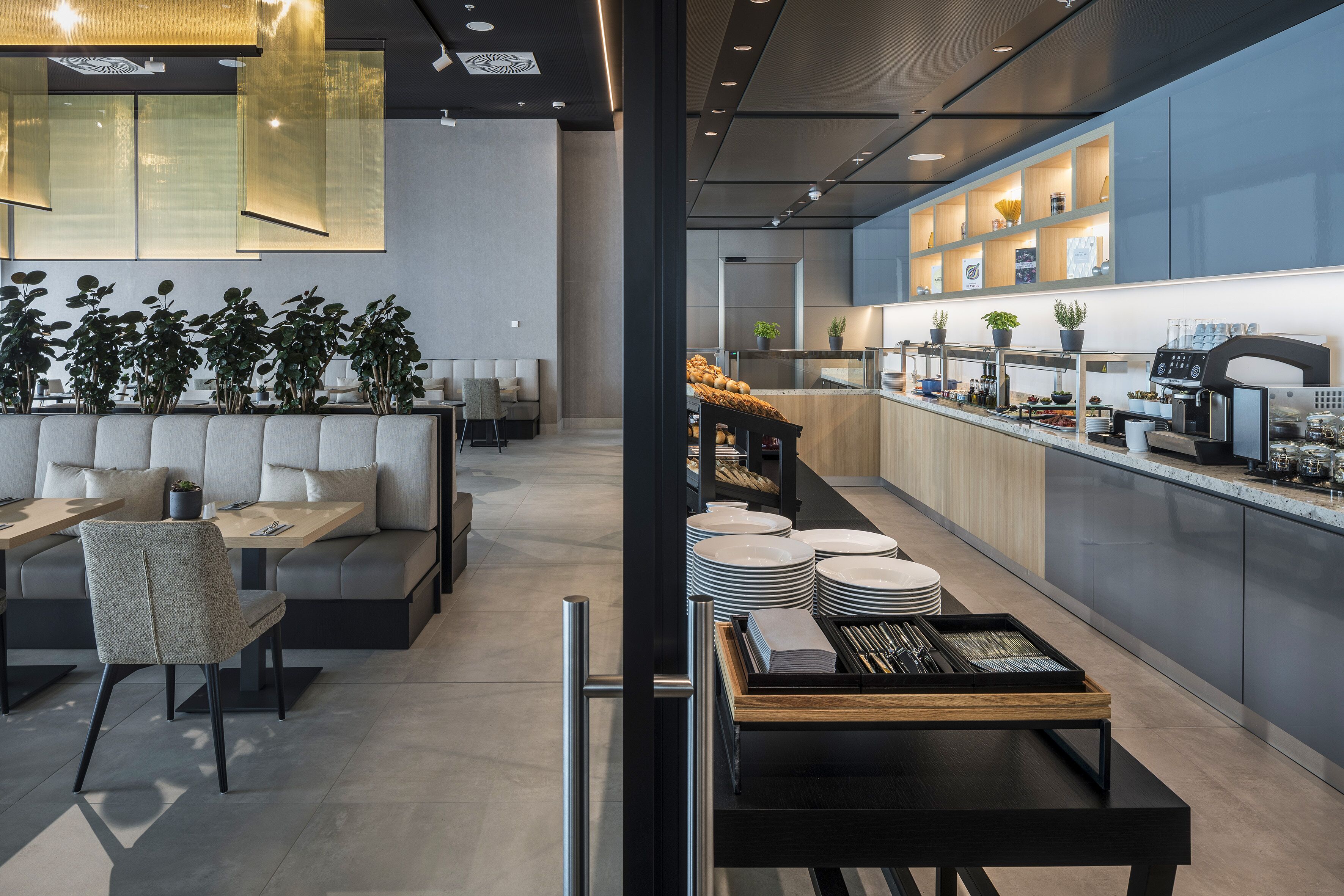 A modern restaurant with a long counter, seating area with cushioned benches, and a kitchen visible through a glass partition.
